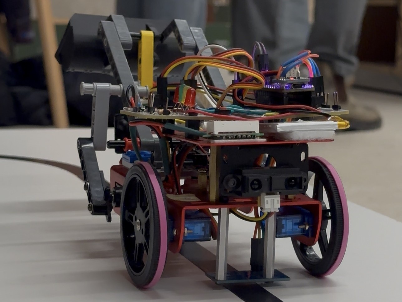 Front view of the skidsteer robot with its red chassis, pink-rimmed wheels and claw arm, following a black line on a white track